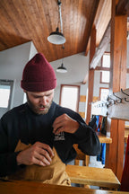 Man wearing a maroon beanie and black sweatshirt with HIGH CAMPBELL text sitting at a wooden table indoors.