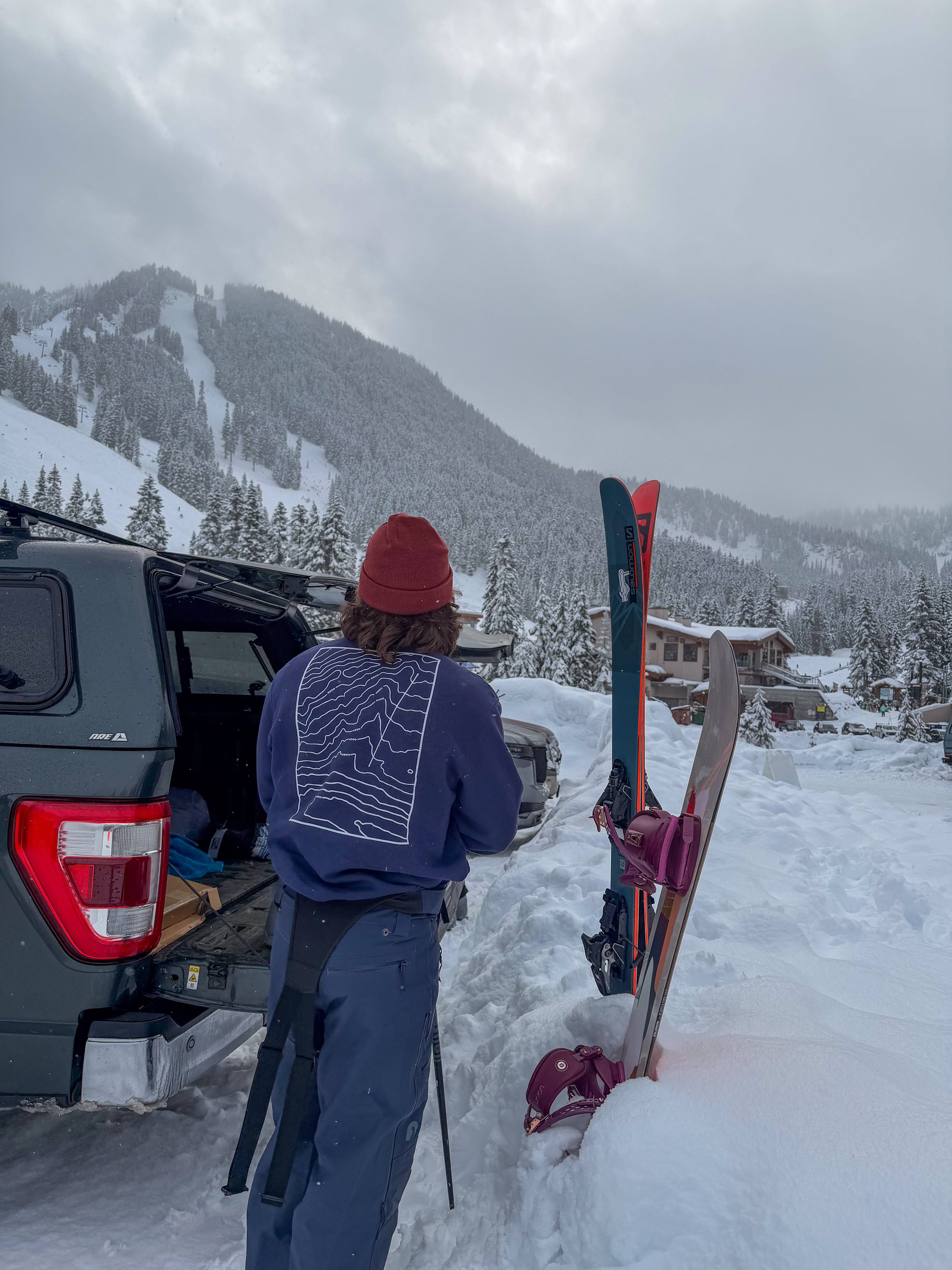 Person with skis preparing to ski in a snowy mountain landscape, navy sweater with white topographic design on back
