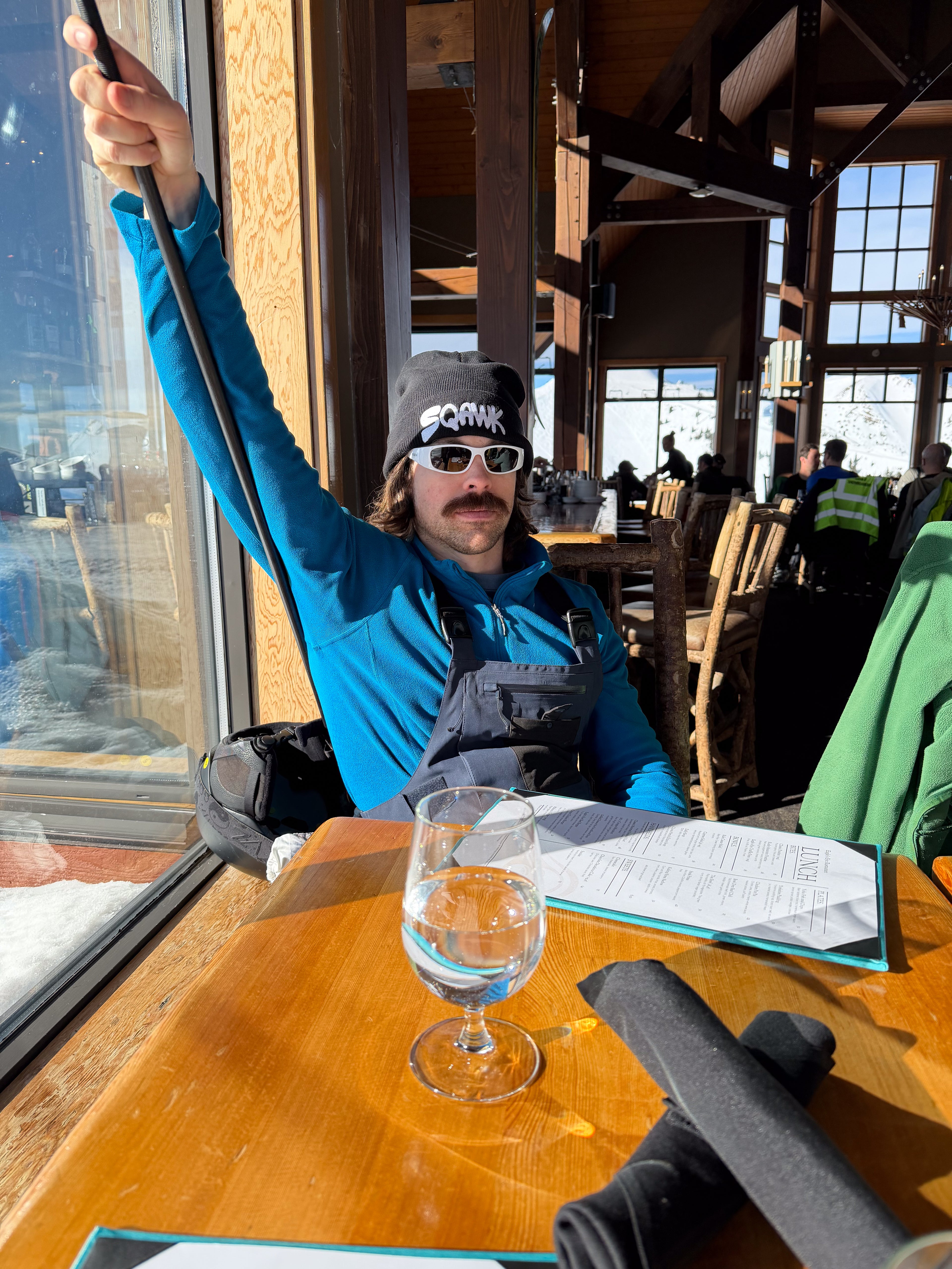 Person sitting at a table with a black SQAWK beanie wearing sunglasses and a blue jacket, in a restaurant setting.